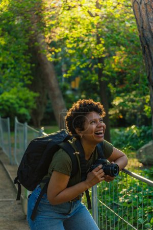 Téléchargez les photos : Jeune photojournaliste afro-américaine noire aux cheveux bouclés foncés riant joyeusement en regardant quelque chose de merveilleux à venir avec un appareil photo à la main au coucher du soleil avec le soleil derrière le rétroéclairage - en image libre de droit