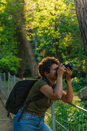 Téléchargez les photos : Jeune photojournaliste afro-américaine noire aux cheveux bouclés foncés prenant des photos dans un parc vert au coucher du soleil avec le soleil couchant derrière elle en ajustant la bague de mise au point de l'appareil photo - en image libre de droit