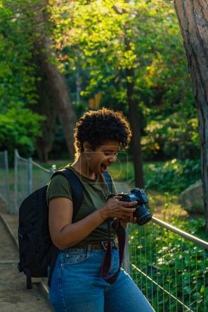 Téléchargez les photos : Heureux jeune noir afro-américain photojournaliste avec sombre cheveux bouclés vérification étonné et heureux avec bouche ouverte le résultat de photos prises avec un appareil photo numérique sur écran numérique dans le parc vert au coucher du soleil avec des paysages - en image libre de droit