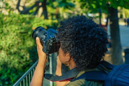Téléchargez les photos : Jeune afro-américaine noire avec des cheveux bouclés foncés photojournaliste prenant une photo dans un parc vert en ajustant la bague de mise au point et les paramètres de l'appareil photo - en image libre de droit