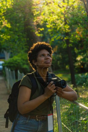 Téléchargez les photos : Jeune photojournaliste afro-américaine noire aux cheveux bouclés foncés à la recherche d'oiseaux à photographier avec appareil photo à la main avec le soleil rétro-éclairé derrière - en image libre de droit