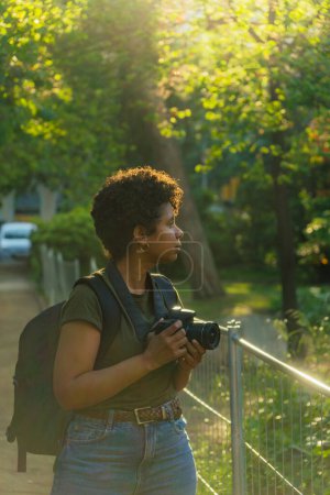 Téléchargez les photos : Jeune photojournaliste afro-américaine noire aux cheveux bouclés foncés à la recherche de quelque chose de merveilleux à photographier avec appareil photo à la main avec le soleil rétroéclairé derrière - en image libre de droit