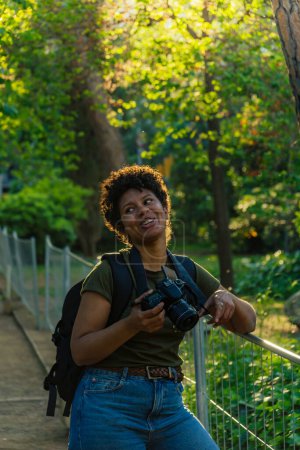 Téléchargez les photos : Heureuse jeune femme noire afro-américaine photojournaliste aux cheveux bouclés foncés riant joyeusement en regardant un ami avec appareil photo accroché au cou dans un parc vert au coucher du soleil avec le soleil derrière backlightin - en image libre de droit