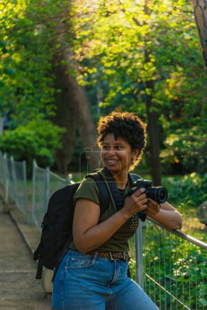 Téléchargez les photos : Jeune photojournaliste afro-américaine noire aux cheveux bouclés foncés riant joyeusement en regardant quelque chose de merveilleux à photographier avec un appareil photo à la main avec le soleil rétroéclairé derrière - en image libre de droit