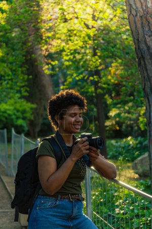 Téléchargez les photos : Jeune photojournaliste afro-américaine noire aux cheveux bouclés foncés et aux yeux fermés souhaitant quelque chose de merveilleux à photographier avec un appareil photo à la main avec le soleil rétroéclairé derrière - en image libre de droit