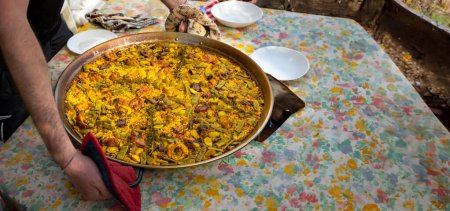 Chef holding a paella pan with a freshly made authentic Valencian paella, to serve on a table with the dishes set and the tablecloth of a table in a rustic farmhouse