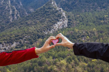 Téléchargez les photos : Deux mains touchant la forme d'un cœur et tenant une figure d'un cœur rose avec des bords rouges avec un fond montagneux luxuriant de haute montagne - en image libre de droit