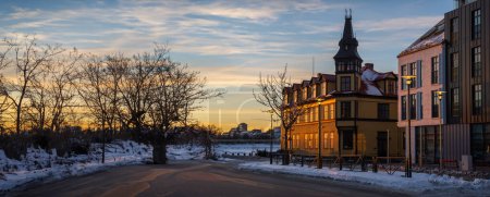 Téléchargez les photos : Facade of typical Reykjavik houses illuminated by the last rays of the evening sun reflecting orange and gold light with snowy sidewalks and a subtly cloudy sky with twilight tones - en image libre de droit