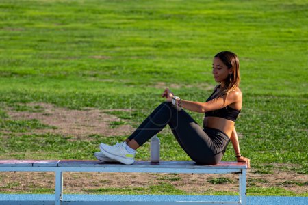 Photo for Beautiful young sporty girl, tanned with long dark hair, sitting on a wooden bench, checking her heart rate with her smart watch after her running workout, the evening sun illuminating her tanned back - Royalty Free Image