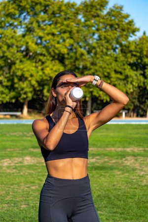 Photo for Beautiful young runner, tanned with long dark hair, protecting her eyes from the sun with her hand, looking at the horizon and drinking and hydrating herself with a can of isotonic drink. - Royalty Free Image