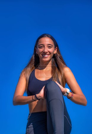 Photo for Beautiful young, tanned woman with long dark hair, smiling and stretching her legs after a hard running workout. Looking at camera happy with a clear blue sky. - Royalty Free Image