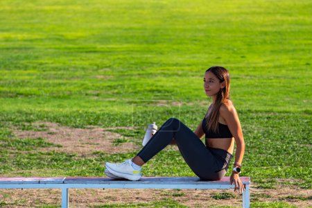 Photo for Beautiful young girl dressed in tight sports clothing, tanned with long hair, sitting on a wooden bench, looking intently at the horizon concentrating before a running race. - Royalty Free Image