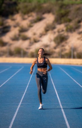 Beautiful slim tanned runner girl, dressed in tight sportswear, smiling happily and enjoying the sun on her face running energetically head-on on a blue running track.