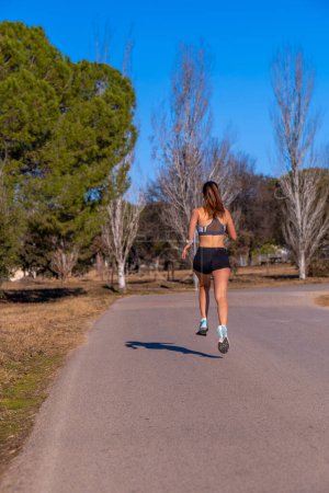 Photo for Young muscular female runner, dressed in tight shorts and top, mid-air running on an asphalt path through a mountain park illuminated by dawn sunlight casting her shadow. - Royalty Free Image