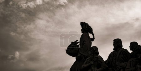 Hinterleuchtete Silhouetten von Kalksteinskulpturen und Heinrich der Seefahrer hält in der Hand ein Schiff vom Denkmal der Entdeckungen in Lissabon mit spektakulärem, bewölkten Himmel bei Sonnenuntergang.