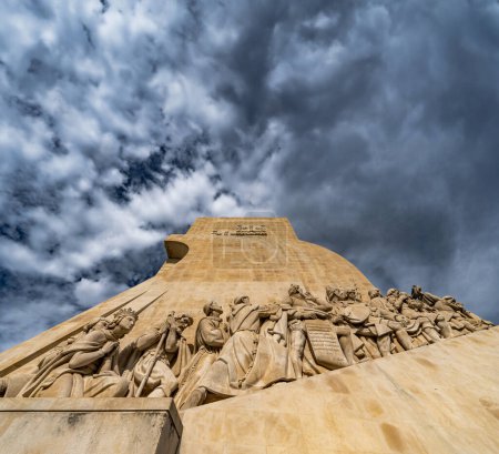Foto de Vista de bajo ángulo del lado occidental de las esculturas de piedra caliza de los navegantes pioneros en el Monumento a los Descubrimientos en Lisboa, Portugal, bajo un cielo azul nublado dramático. - Imagen libre de derechos