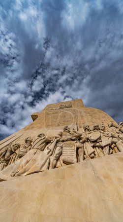 Tiefansicht des westlichen Profils mit Kalksteinskulpturen von Pioniernavigatoren vom Monument der Entdeckungen in Lissabon, Portugal, unter einem dramatisch bewölkten blauen Himmel. Vertikales Banner.