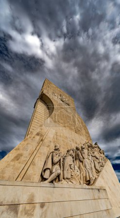 Low angle view of the western skyline with limestone sculptures of pioneer navigators from the Monument to the Discoveries in Lisbon, Portugal, under a dramatic cloudy blue sky. Vertical flag.