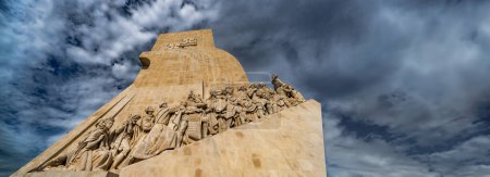 View from below of the western profile with limestone sculptures and Henry the Navigator on the bow of the Monument to the Discoveries in Lisbon, Portugal, under a cloudy blue sky. Horizontal Banner.