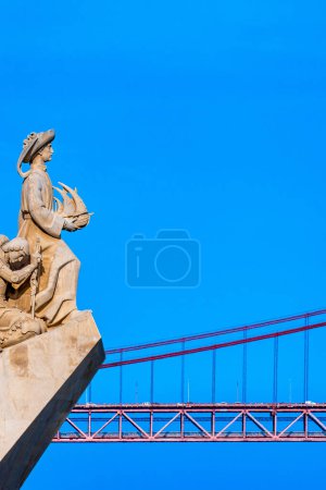 Western profile of the Monument to the Discoveries, with Henry the Navigator holding a boat looking at the red steel 25 de Abril suspension bridge. Clear blue sky.