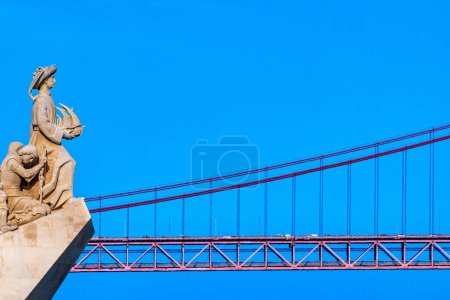 Foto de Perfil occidental del Monumento a los Descubrimientos, con Enrique el Navegante sosteniendo un barco mirando el puente colgante de acero rojo 25 de Abril. Cielo azul claro. - Imagen libre de derechos