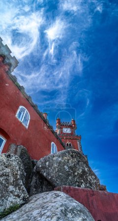 Vue en angle bas d'une partie d'un mur rouge du palais de Pena sur de grands rochers sous un ciel bleu avec des nuages de toile d'araignée blanche rétroéclairés par le soleil. Sintra. Portugal.