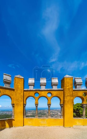 Moorish arches of the arched courtyard with the yellow painted walls of the Pena Palace, with the Sintra mountain range and the Atlantic Ocean on the horizon, under a clear blue sky.