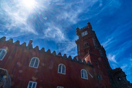 Photo for Low angle view of the red facade showing the silhouette of the Pena Palace with the clock tower under a blue sky with clouds in white cobwebs backlit by the sun. Sintra. Portugal. - Royalty Free Image