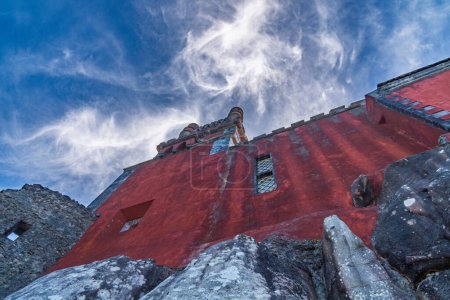 Niedrige Ansicht eines Teils einer roten Wand des Pena-Palastes auf einigen großen Felsen unter blauem Himmel mit weißen Spinnwebenwolken, die von der Sonne hinterleuchtet werden. Sintra. Portugal.