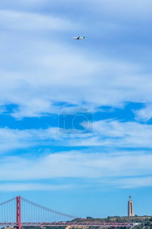 Foto de Avión comercial con turistas volando sobre el puente colgante de acero rojo 25 de Abril y el Sagrado Corazón de Jesús de Almada monumento religioso en Lisboa. - Imagen libre de derechos