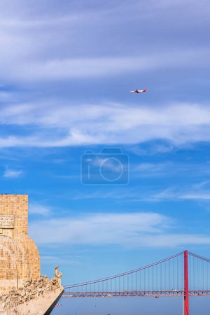 Western profile of the Monument to the Discoveries, with tourists on the attic looking at the bridge 25 de Abril and a commercial airliner flying through the sky.