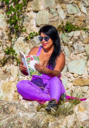 Sexy dunkelhäutige Latina-Frau trägt rosa enge Leggings und Top ein Buch im Freien in der Landschaft lächelnd glücklich und fröhlich aussehend mit ländlichem Hintergrund.