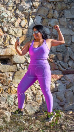 Sexy übergewichtige Latina mit dunkler Haut, gekleidet in enge rosa Sportbekleidung, mit der Faust in einem Siegessymbol, die glücklich auf ihr Smartphone blickt, nachdem sie ihre Sportergebnisse gesehen hat.
