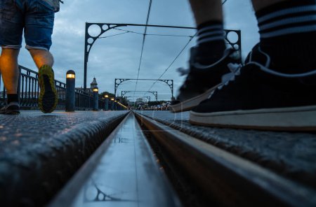 Close-up of men's feet with sneakers and shorts crossing the Porto metro track on the Dom Luis bridge at sunset, with the reflection of the cables and arches on the train track.