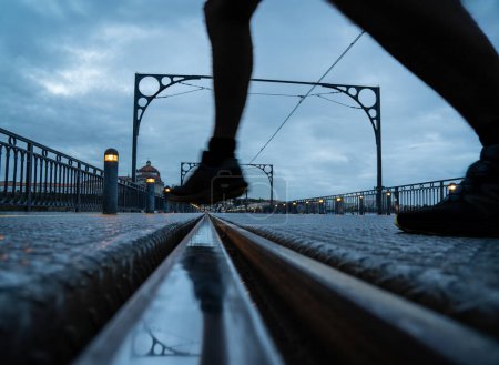 Silhouette of man's legs taking a step to cross the Porto metro track on the Dom Luis bridge at sunset, with the reflection of the cables, arches and the leg on the train track.