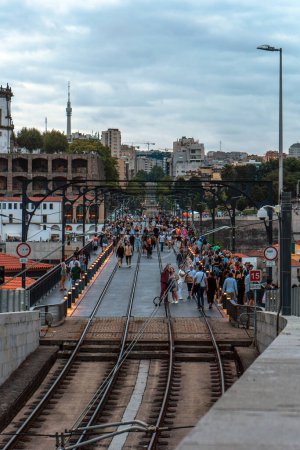 Foto de Vista panorámica de la vía del metro de Oporto desde el puente Dom Luis al atardecer, con las luces en los polos llenos de turistas caminando bajo un cielo nublado. - Imagen libre de derechos