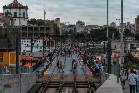 Foto de Vista panorámica de la vía del metro de Oporto desde el puente Dom Luis al atardecer, con las luces en los polos llenos de turistas caminando bajo un cielo nublado. - Imagen libre de derechos