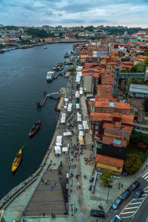 Photo for Aerial drone view of the Douro river and the Porto seafront with the old houses, and classic boats at the dock; restaurants and tourists strolling along the river walk at sunset under a cloudy sky. - Royalty Free Image