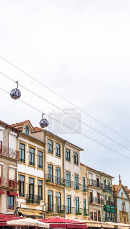 Two gondolas with tourists from the Gaia cable car passing just above the typical houses of the Vila Nova de Gaia neighborhood and by the umbrellas of the restaurants, under a cloudy sky in Porto.