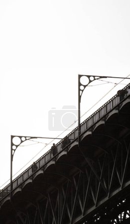 Silhouette von Menschen, die auf der oberen Plattform der Stahlbrücke Don Luis I in Porto gehen, mit Details ihrer Metallstruktur und den Kabeln, durch die die U-Bahn von Porto unter weißem Himmel fährt.