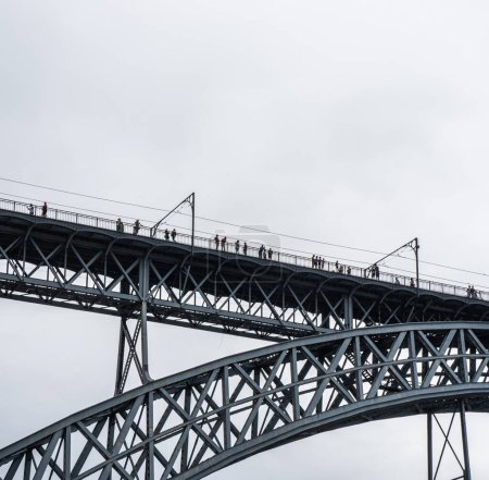 Foto de Turistas caminando, tomando fotos y una mujer posando desde la plataforma superior del puente de acero Don Luis I donde pasa el metro de Oporto con nubes de lluvia. - Imagen libre de derechos