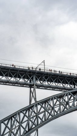 Téléchargez les photos : Touristes marchant, prenant des photos et une femme posant depuis la plate-forme supérieure du pont en acier Don Luis I où passe le métro de Porto avec des nuages de pluie. - en image libre de droit