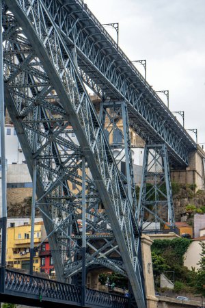 Vista de bajo ángulo del puente de acero Don Luis I en Oporto y personas caminando y tomando fotos del río Duero en la plataforma superior con casas del antiguo barrio de abajo.