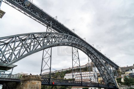 Vista de perfil bajo del puente de acero Don Luis I en Oporto con nubes de lluvia en el fondo y gente caminando y tomando fotos del río Duero en la plataforma superior y gente en restaurantes.