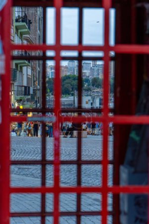 Calle turística Cobblestone en Oporto al atardecer, llena de turistas caminando con cielo nublado visto a través de una típica cabina telefónica roja portuguesa.
