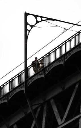 Téléchargez les photos : Couple d'amoureux marchant le long du pont en acier Don Luis I à Porto avec des détails de sa structure métallique avec des ornements sous un ciel blanc nuageux. - en image libre de droit