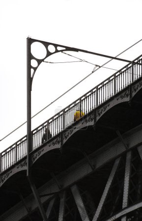 Téléchargez les photos : Homme en t-shirt jaune et femme vêtue de noir aux cheveux longs, marchant sur le pont en acier Don Luis I à Porto avec des détails de sa structure métallique avec des ornements sous un ciel blanc nuageux. - en image libre de droit
