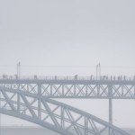 People with umbrellas and raincoats walking on the upper platform of the Don Luis I steel bridge and cars driving on the lower platform, with a person running surrounded by fog and rain clouds.