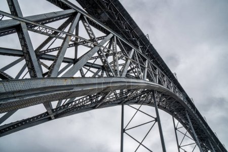 Vista de perspectiva con detalles de remaches y estructura metálica desde abajo del puente de acero Don Luis I en Oporto con nubes de lluvia en el fondo.