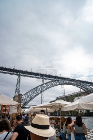 Téléchargez les photos : Les gens et les touristes faisant du shopping au marché de Ribeira, sur la promenade Vila de Gaia à Porto, avec le pont Dom Luis I en arrière-plan avec des piétons marchant sous un ciel nuageux. - en image libre de droit